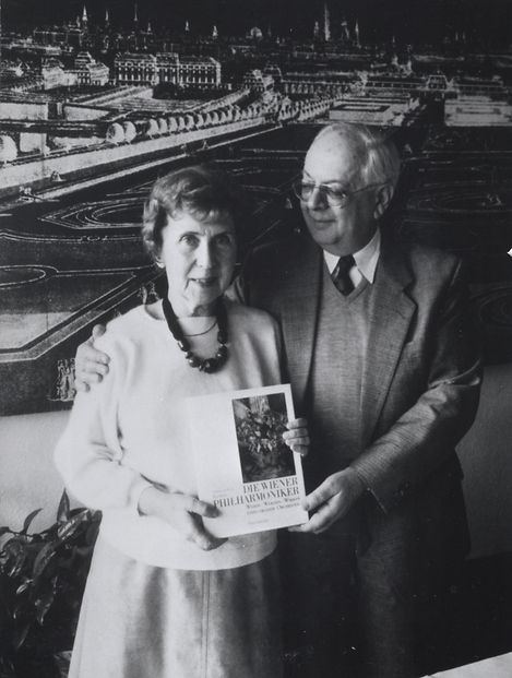 You see a black and white photograph of Herta and Kurt Blaukopf presenting their book "The Vienna Philharmonic" to the camera. Herta Blaukopf is looking straight into the camera, while Kurt Blaukopf has his arm around her and is looking at his wife.